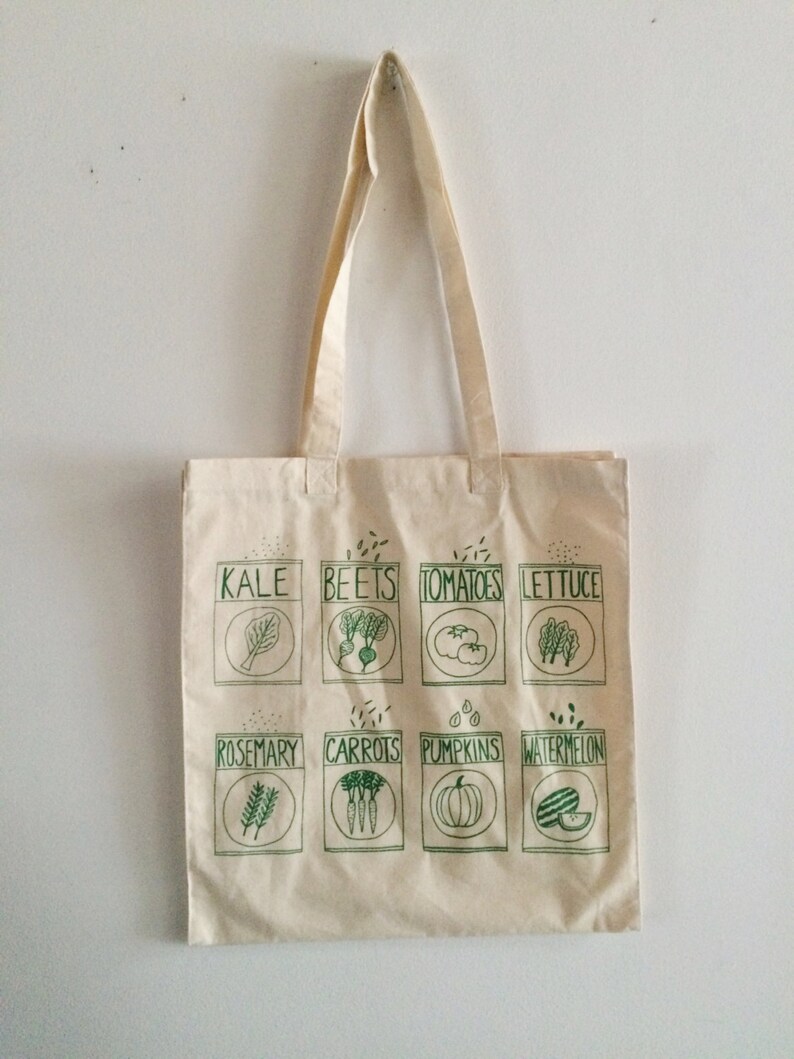 May include: A white canvas tote bag with green illustrations of various vegetables and herbs. The bag has two long handles and features the words "Kale", "Beets", "Tomatoes", "Lettuce", "Rosemary", "Carrots", "Pumpkins", and "Watermelon" printed on it.
