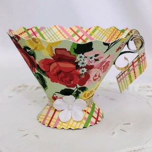 May include: A paper teacup with a floral pattern and a plaid ribbon around the base. The cup is decorated with a white flower and a gold ribbon.