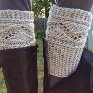 May include: A pair of beige knitted leg warmers with a cable knit pattern. The leg warmers are worn over black boots.