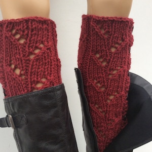 May include: A pair of red knitted leg warmers with a lacy pattern. The leg warmers are worn over black boots.