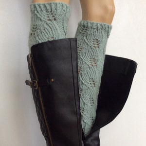 May include: A pair of light green knitted leg warmers with a lacy pattern. The leg warmers are worn over black leather boots with a zipper.