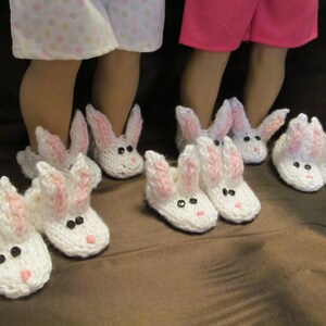 May include: A pair of white knitted bunny slippers with pink ears. The slippers have black button eyes and are worn by two dolls.