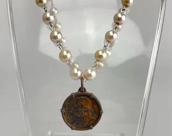 May include: A silver chain necklace with a round, brown, metal pendant. The necklace is made of alternating cream and light brown pearls.