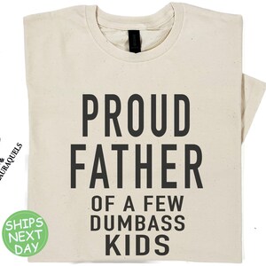 May include: A natural t-shirt with the text 'PROUD FATHER OF A FEW  DUMBASS KIDS' printed in black.