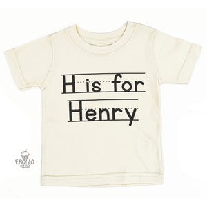 May include: Cream-colored short-sleeved t-shirt with the text "H is for Henry" in black, designed to look like handwriting on lined paper. The shirt is a children's apparel item.