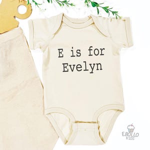 May include: Cream-colored baby onesie with the text "E is for Evelyn" in black. A light-colored tote bag and a wooden cutout are next to the onesie. Greenery is at the top. The logo "EBOLLO KIDS" is at the bottom.