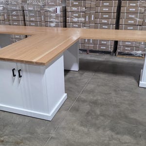 May include: L-shaped wooden desk with a light brown tabletop and white base. The desk has drawers and cabinet doors with black handles. The desk is in a warehouse setting.