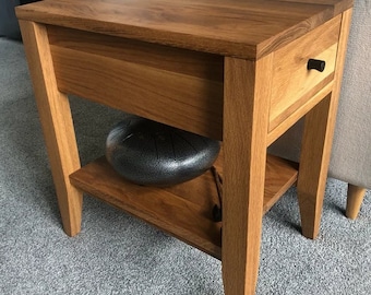 May include: A wooden side table with a drawer and a lower shelf. The table is made of light brown wood and has a black drawer pull. A gray metal instrument sits on the lower shelf. The table is next to a gray sofa.