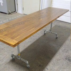 May include: A long, rectangular wooden table with a natural wood finish. The table is supported by a metal pipe frame with wheels. The table is in a warehouse setting.
