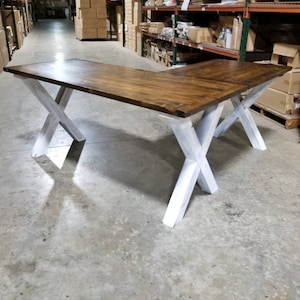 May include: L-shaped wooden desk with a dark brown tabletop and white X-shaped legs. The desk is made of wood and has a natural finish. The desk is in a warehouse setting.