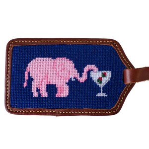 Può includere: Un elefante rosa con la proboscide avvolta attorno a un bicchiere di bacche rosse. L'elefante e il bicchiere sono ricamati su uno sfondo blu. Lo sfondo è circondato da un bordo in pelle marrone. Il cinturino in pelle è attaccato alla parte superiore del bordo.