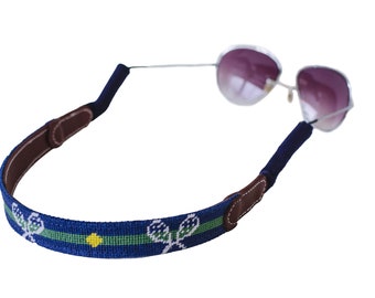 May include: A blue and green patterned eyeglass strap with a brown leather accent. The strap features a tennis racket design.