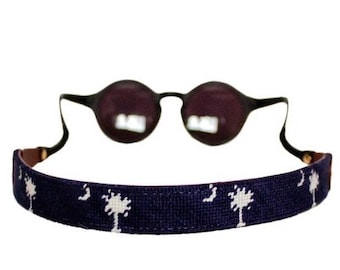May include: Round sunglasses with dark lenses and a navy blue strap featuring a white palm tree and crescent moon pattern. The strap is attached to the glasses' arms.