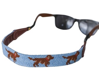 May include: Sunglasses with a light blue strap featuring a repeating pattern of brown dogs with red collars. The strap is attached to the sunglasses with brown leather and black arms.