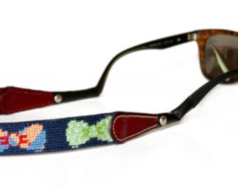 May include: Sunglasses with a navy blue needlepoint strap featuring a pattern of bow ties and other shapes in blue, peach, and green. The strap has dark red leather accents and black leather ends that attach to the sunglasses.