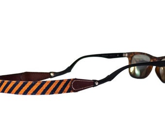 May include: Sunglasses with a black frame and brown lenses are attached to a striped strap. The strap features a pattern of orange and navy blue stripes. The strap is made of fabric and leather.