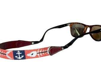 May include: Sunglasses with a beaded strap featuring a nautical design. The strap is coral with white and navy blue accents, including an anchor motif. The sunglasses have a tortoiseshell frame and black arms.