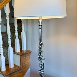 May include: A unique lamp featuring a silver clarinet as the central support. The lamp has a white lampshade with a light blue trim and sits on a black base. The lamp is placed on a wooden table.