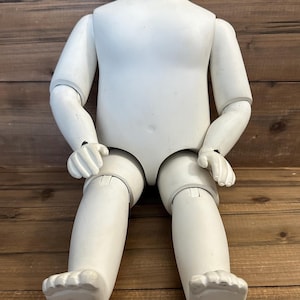 May include: A white, articulated doll body with a rounded neck, arms, and legs. The doll's limbs are jointed, allowing for posing. The doll is made of a solid, smooth material and is resting on a wooden surface.