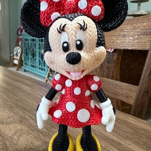 Pu&ograve; includere: Una statuina di Minnie Mouse stampata in 3D. La figura presenta un vestito rosso a pois bianchi, un fiocco abbinato e scarpe gialle. Ha orecchie, braccia e gambe nere e guanti bianchi. La statuina &egrave; su una superficie di legno.