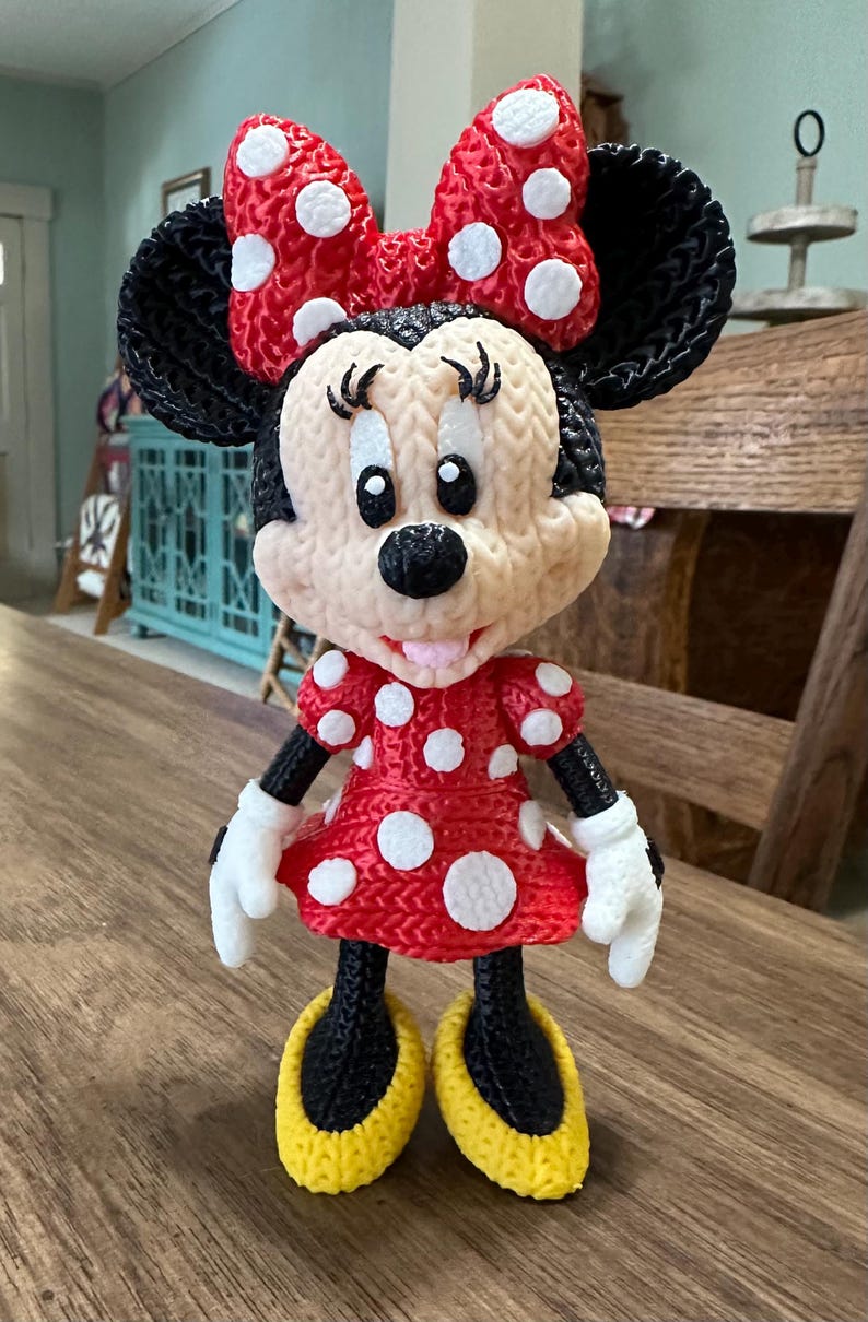 Pu&ograve; includere: Una statuina di Minnie Mouse stampata in 3D. La figura presenta un vestito rosso a pois bianchi, un fiocco abbinato e scarpe gialle. Ha orecchie, braccia e gambe nere e guanti bianchi. La statuina &egrave; su una superficie di legno.