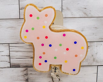 Könnte beinhalten: Rosa-goldener Glitzer-Badgehalter in Form eines Zuckerkekses mit bunten Streuseln. Der Halter hat eine einziehbare Schnur und einen Clip.