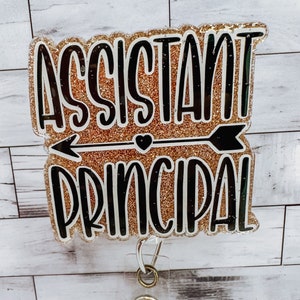 May include: A gold glitter background with black text that reads "Assistant Principal" with a black arrow and heart in the middle. A clear badge holder with a retractable cord is attached to the bottom.
