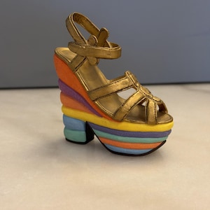 May include: A gold-colored platform sandal with a rainbow-colored wedge heel. The sandal features a strap around the ankle and a strap across the toes. The heel is composed of layers of orange, purple, yellow, and blue.