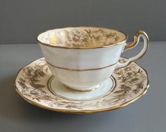 Può includere: Un set di tazza da tè e piattino in porcellana bianca con un delicato motivo floreale dorato e bordi smerlati rifiniti in oro. La tazza presenta un manico curvo e una banda dorata orizzontale, mentre il piattino mostra un motivo floreale coordinato.