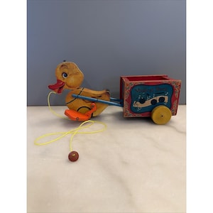 May include: Vintage wooden pull-along toy duck with a red cart. The duck is yellow with a red beak and orange feet. The cart is red with a blue musical note design and the words "Musical Duck". The toy has yellow string and wooden wheels.