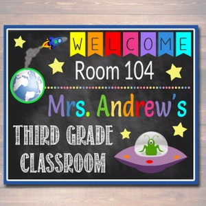 Puede incluir: Un cartel de bienvenida para el aula en una pizarra con tem&aacute;tica espacial. Dice "Bienvenido" en letras arco&iacute;ris, "Sala 104", "Clase de tercer grado de la Sra. Andrew", y presenta un cohete, estrellas, un planeta Tierra y un platillo volador con un extraterrestre.
