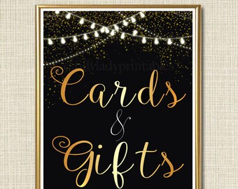 May include: A black and gold sign with the text "Cards & Gifts" in gold script font. The background is black with gold glitter and a string of white lights.