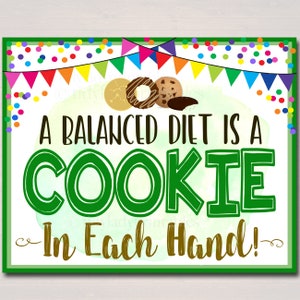 May include: A green and gold sign with confetti and a string of colorful flags. The sign reads "A balanced diet is a cookie in each hand!" with three cookies in the top left corner.