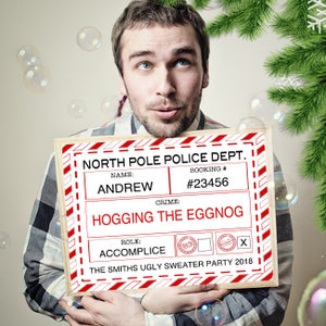 Peut inclure: Un panneau blanc avec des rayures rouges et le texte "NORTH POLE POLICE DEPT."  Le panneau porte le texte "NAME: ANDREW", "BOOKING #: #23456", "CRIME: HOGGING THE EGGNOG", "ROLE: ACCOMPLICE", et "THE SMITHS UGLY SWEATER PARTY 2018".
