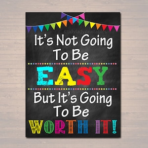 Op de afbeelding: Een zwarte print in schoolbordstijl met de tekst "It's Not Going To Be EASY But It's Going To Be WORTH IT!" in kleurrijke letters. Bovenin staat een banner met kleurrijke vlaggen. De print staat op een houten achtergrond.