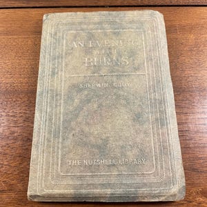 May include: A rectangular book titled An Evening With Burns by Sherwin Cody from The Nutshell Library. The cover is a muted, weathered gray-green with a textured, mottled appearance and features embossed decorative border lines around the edges.