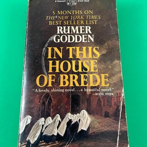 May include: A vintage paperback book cover with a dark brown background. The title "In This House of Brede" is in yellow letters. The author, Rumer Godden, is listed below the title. The cover features a painting of a group of nuns walking in a courtyard.