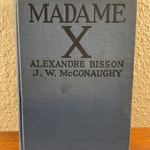 May include: A blue hardcover book titled Madame X by Alexandre Bisson and J. W. McConaughy. The front cover features the title and author names printed in black text against a textured blue fabric background with a small dark spot near the center.