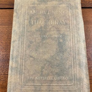 May include: A hardcover book titled An Evening with Thackeray by Sherwin Cody from The Nutshell Library. The cover is a mottled, weathered tan and dark gray color with embossed text and a rectangular border design pressed into the material.