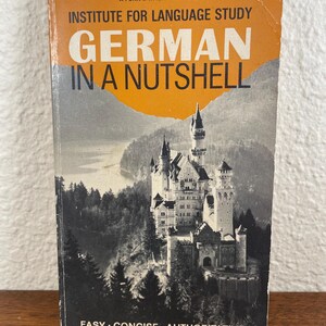May include: A paperback book titled German in a Nutshell by the Institute for Language Study. The cover features a black and white photograph of a castle nestled in a forest near a lake, with orange text and a torn-paper effect at the top.