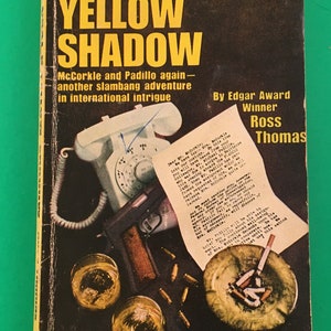 May include: A vintage paperback book cover with a yellow and black title "Cast a Yellow Shadow" by Edgar Award Winner Ross Thomas. The cover features a white rotary phone, a gun, two glasses of whiskey, and an ashtray with cigarettes. The text on the cover reads "Wild, woolly, and as wonderful as THE COLD WAR SWAP." - Library Journal.