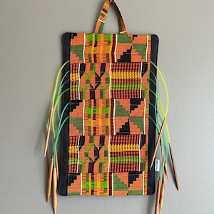 May include: A rectangular knitting needle case with a colorful African print design. The case has a black border and a matching handle. Several knitting needles are visible, with wooden tips and green and yellow cords.