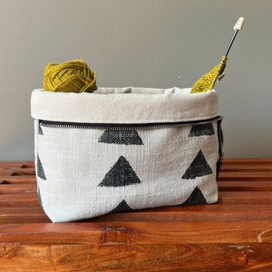 May include: A white canvas storage bag with a black zipper and a black triangle pattern. The bag is filled with a ball of yellow yarn and a knitting needle.