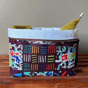 May include: A brown zippered pouch with a white lining and a colorful geometric pattern. The pouch is filled with yarn and a knitting needle.