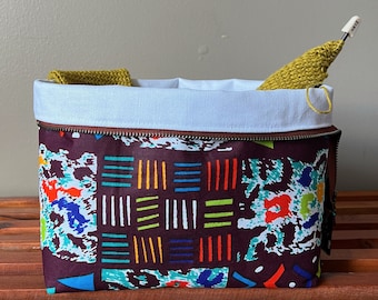 May include: A brown zippered pouch with a white lining and a colorful geometric pattern. The pouch is filled with yarn and a knitting needle.