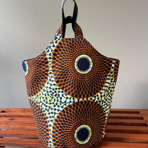 May include: A fabric tote bag with a vibrant African print. The bag features a repeating pattern of orange circles with black dots, yellow and white accents, and a navy blue center. It has black handles and a black interior.