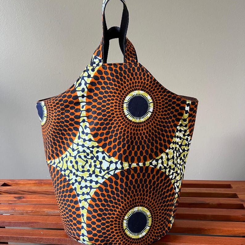 May include: A fabric tote bag with a vibrant African print. The bag features a repeating pattern of orange circles with black dots, yellow and white accents, and a navy blue center. It has black handles and a black interior.