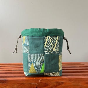 May include: A green patchwork drawstring bag with geometric and abstract patterns in shades of green and yellow. The bag has a green fabric top and a drawstring closure with brown cords. The bag is sitting on a wooden surface.