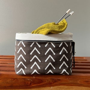 May include: A brown and white fabric knitting project bag with a zipper closure. The bag features a repeating arrow pattern. Inside the bag is a yellow knitted piece with knitting needles.