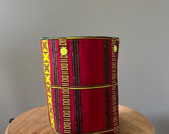 May include: A cylindrical fabric storage bin featuring a vibrant red base with vertical stripes in yellow, black, and green. The exterior is decorated with geometric patterns and includes two bright yellow circular snap buttons near the top rim.